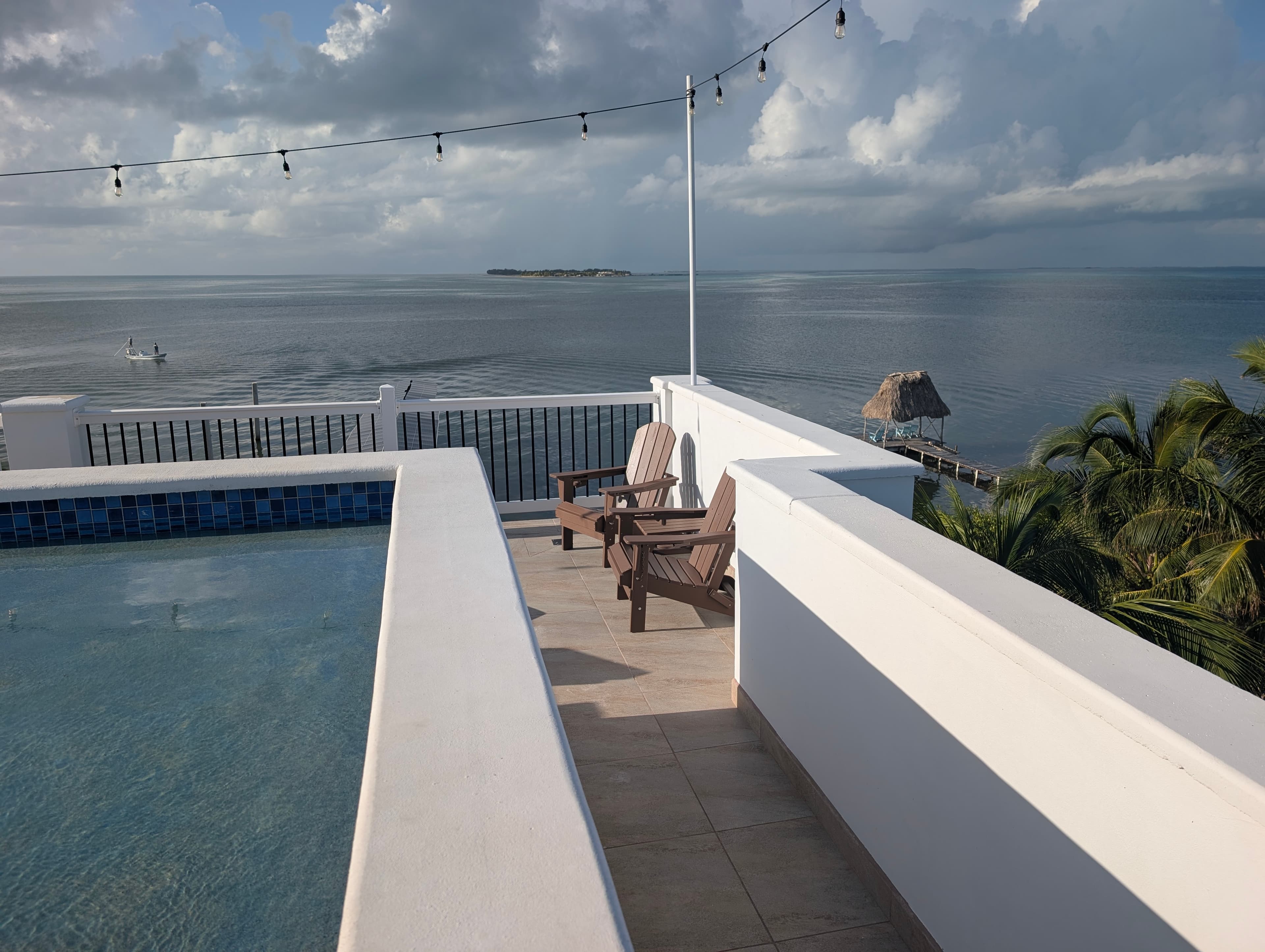 A serene rooftop view featuring a pool, wooden chairs, and the ocean in the background.
