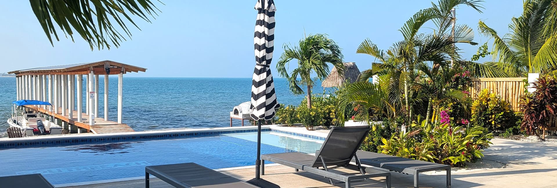 A serene pool area with lounge chairs, a striped umbrella, and a wooden dock overlooking the ocean.