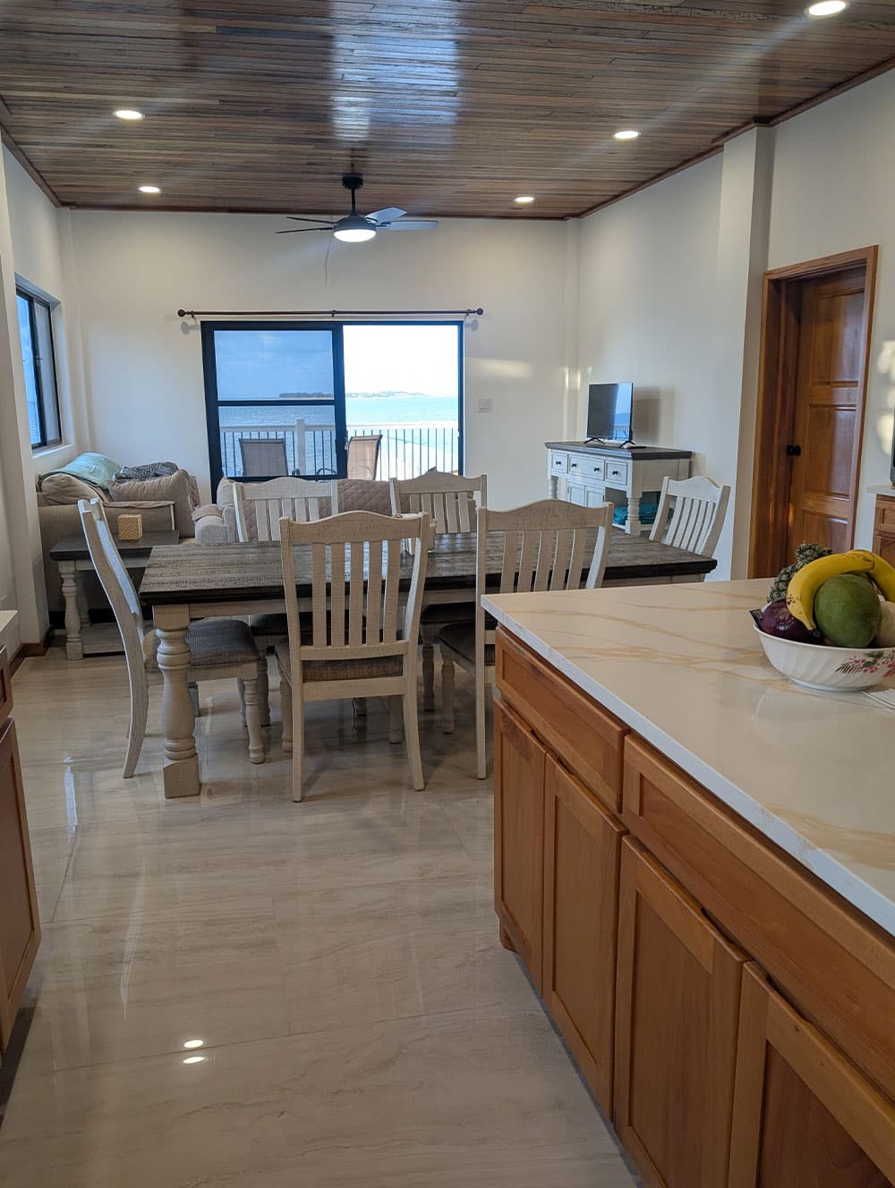 A modern dining area with a large table, wooden accents, and a view of the ocean through a sliding glass door.