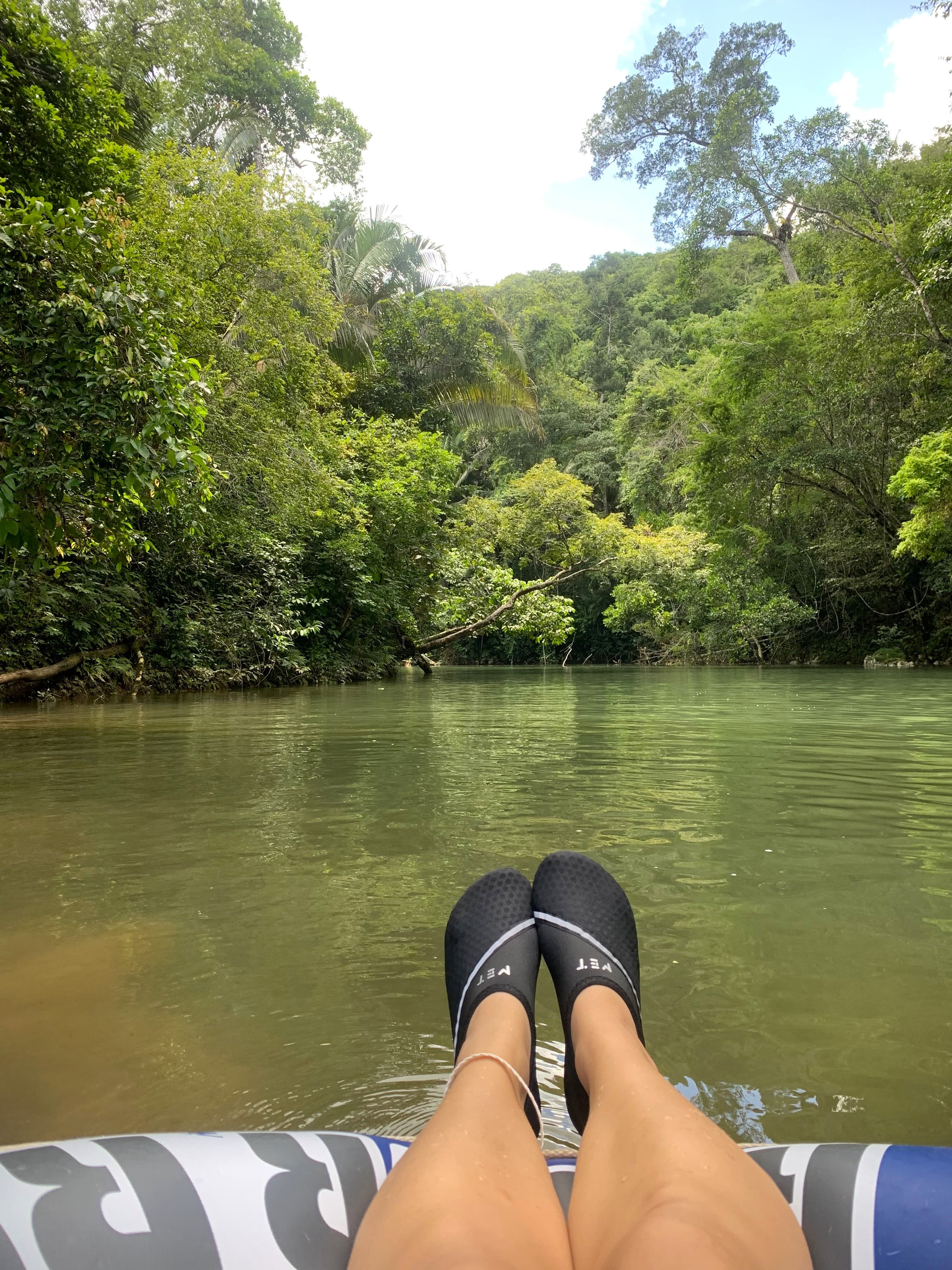 Feet in black water shoes resting on a floating tube in a lush green river setting.