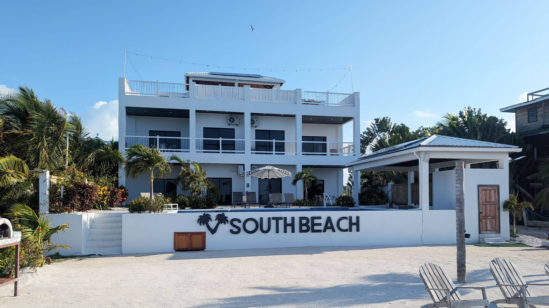 A modern beachfront building with "SOUTH BEACH" signage surrounded by palm trees and a clear blue sky.