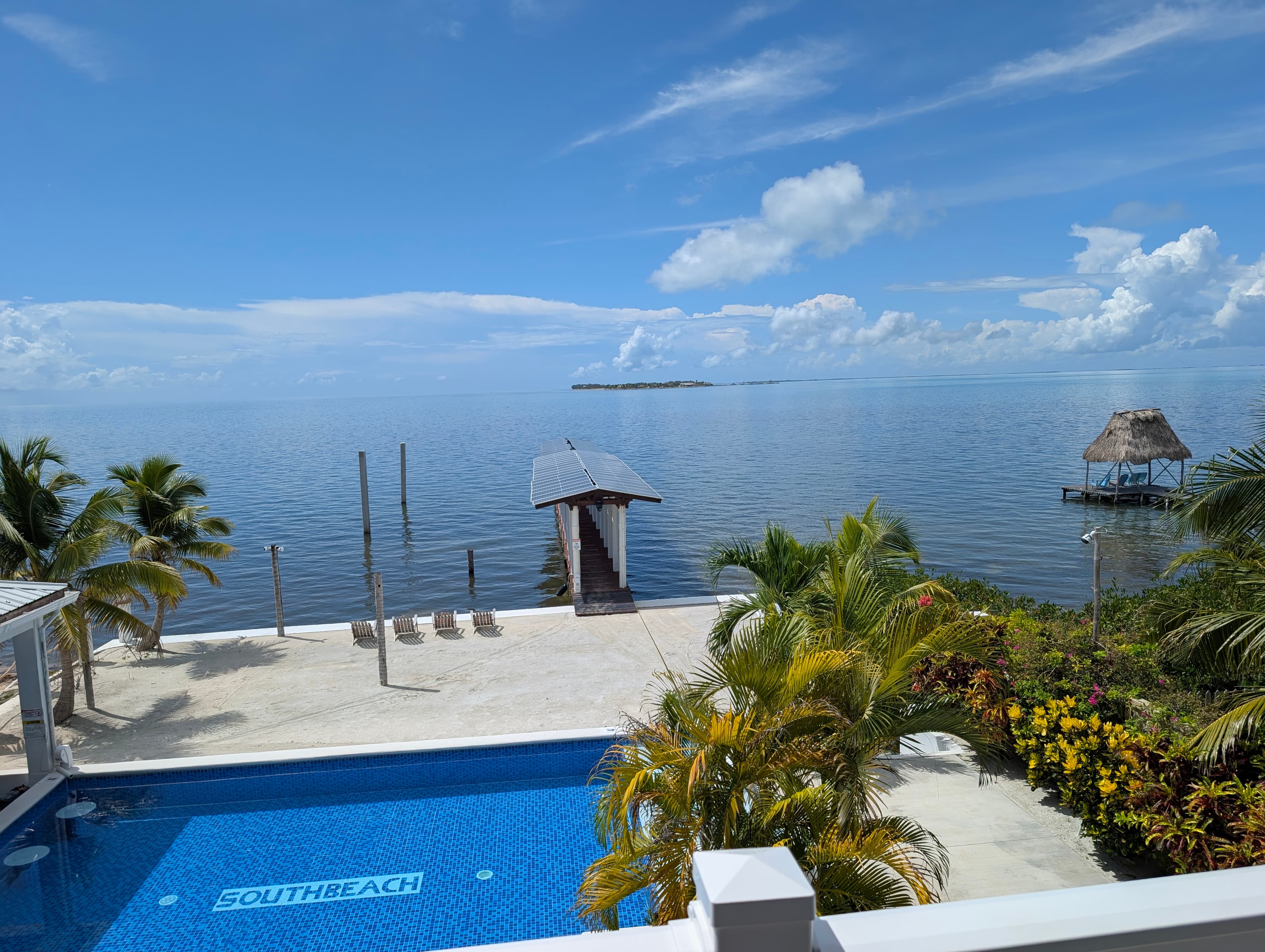A serene view of a tropical beachfront with a swimming pool, palm trees, and a dock extending into the calm waters.