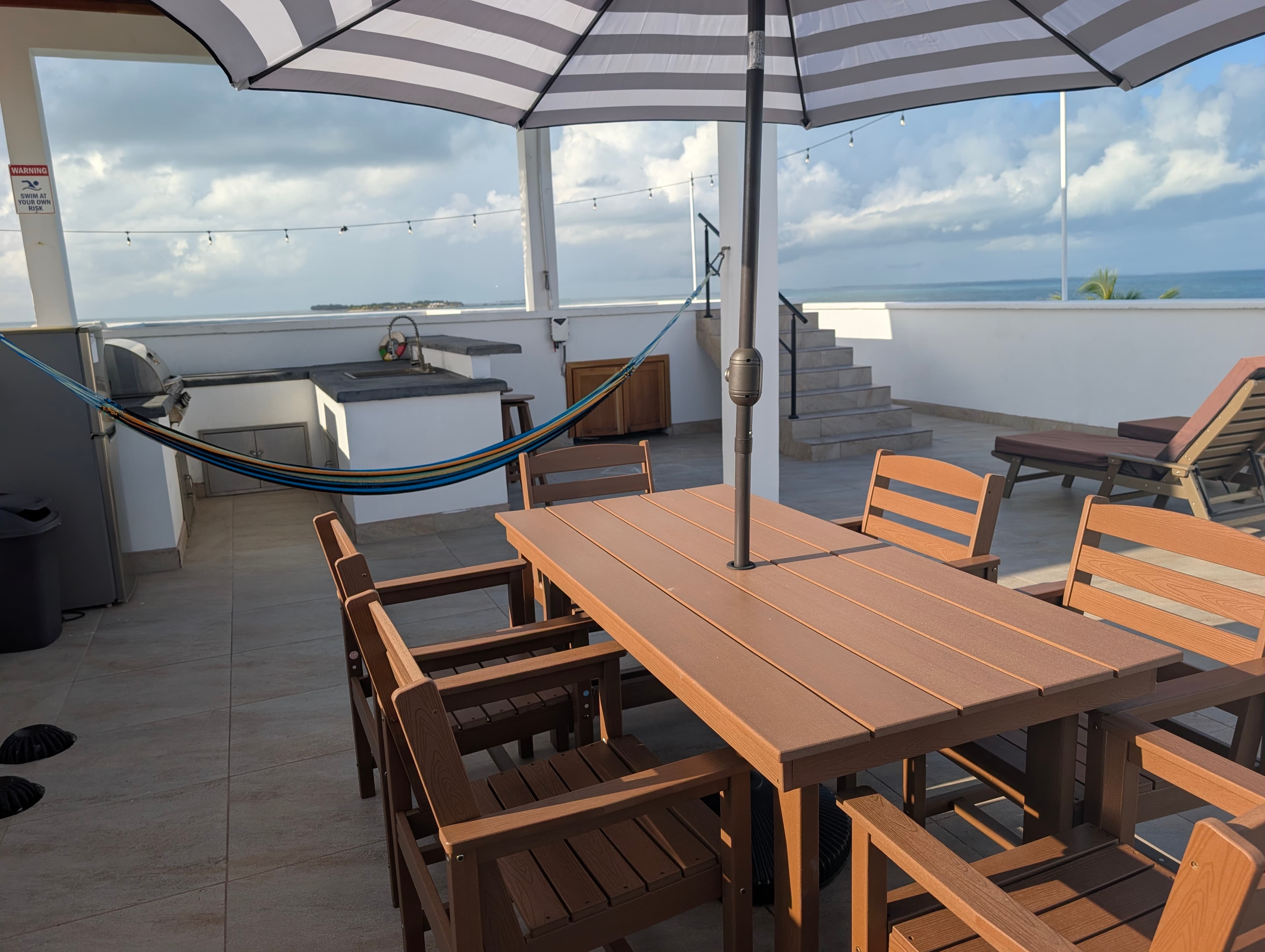 A sunlit terrace featuring a wooden dining table with chairs, an umbrella, a hammock, and a view of the ocean.