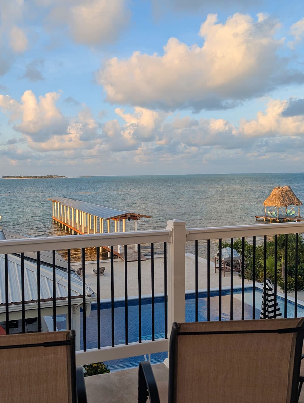 View of a serene waterfront with a pier, thatched gazebo, and swimming pool under a cloudy sky.