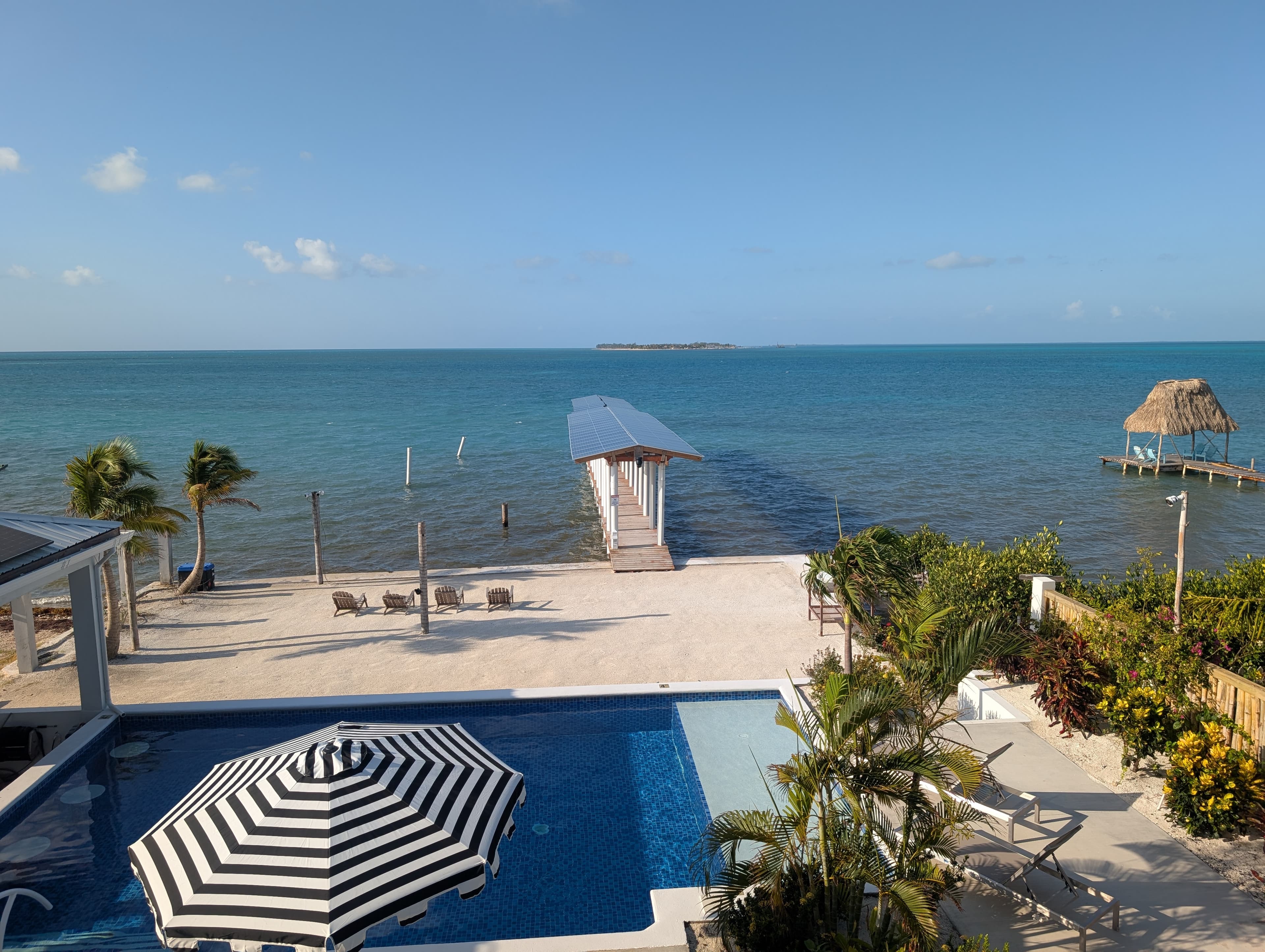 A view of a pool, striped umbrella, and a dock by the turquoise sea under a clear sky.