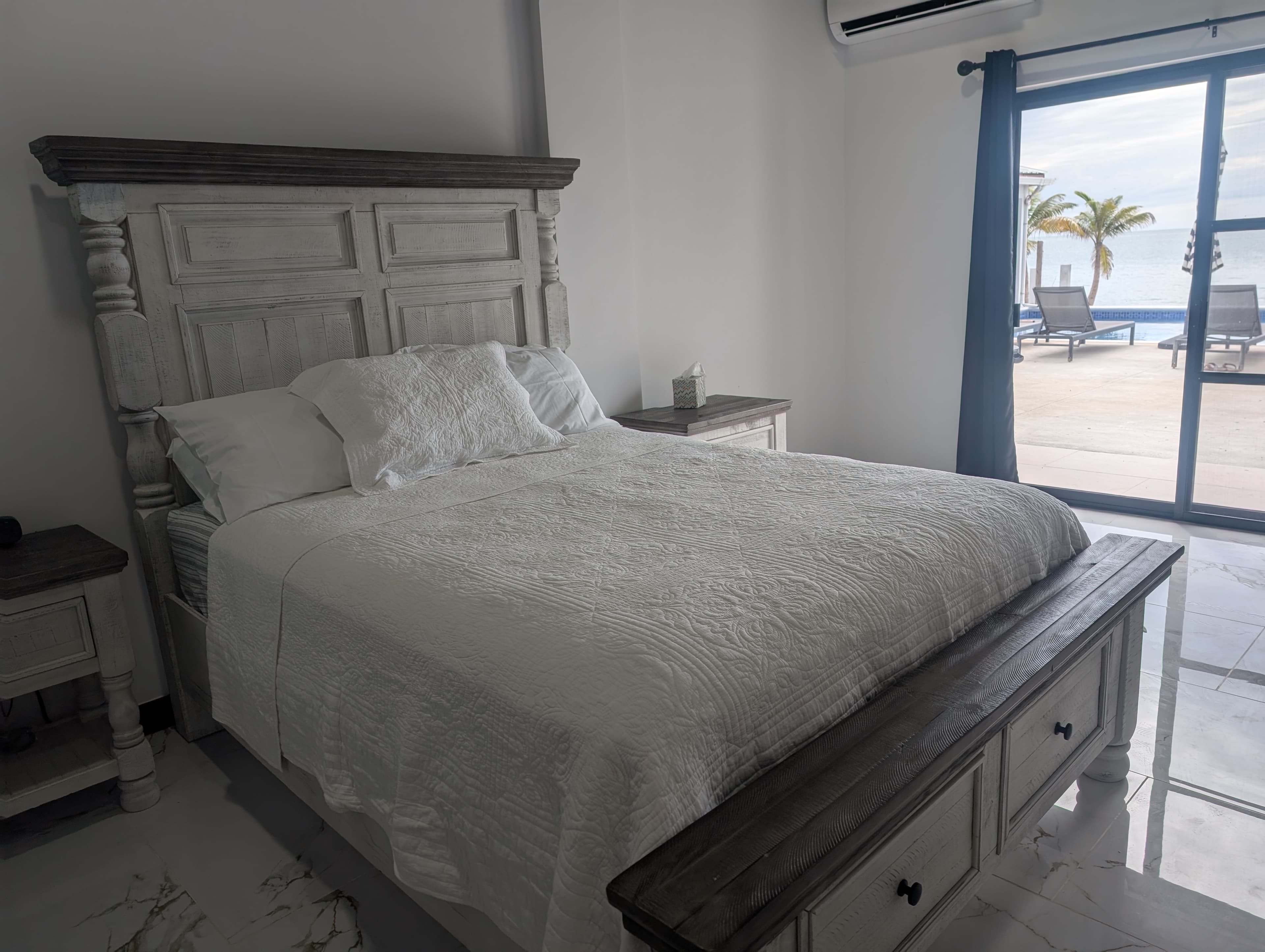 A bright bedroom featuring a white bed with a wooden headboard, beside a glass door leading to an outdoor area with lounge chairs.