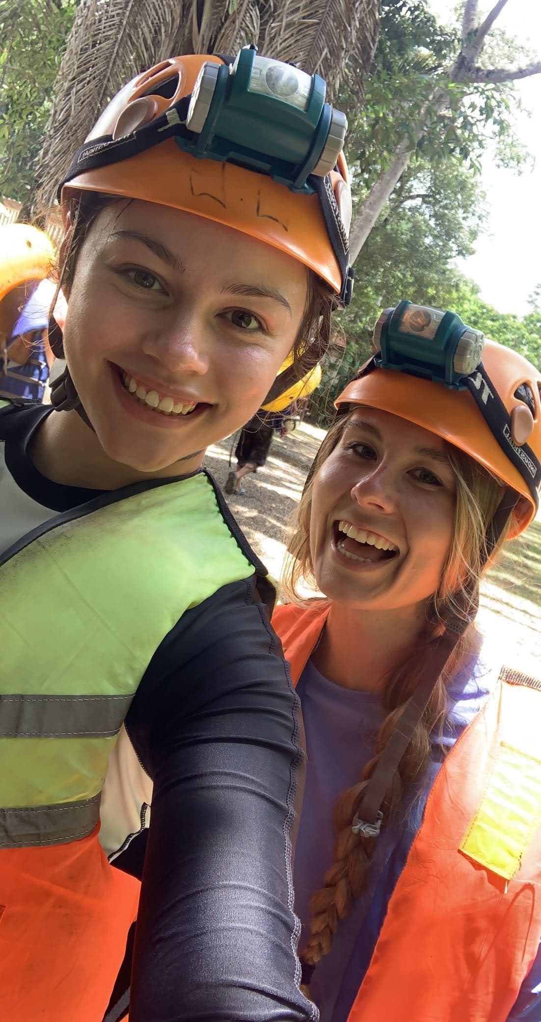Two smiling individuals in helmets and safety vests pose for a selfie in a forested area.