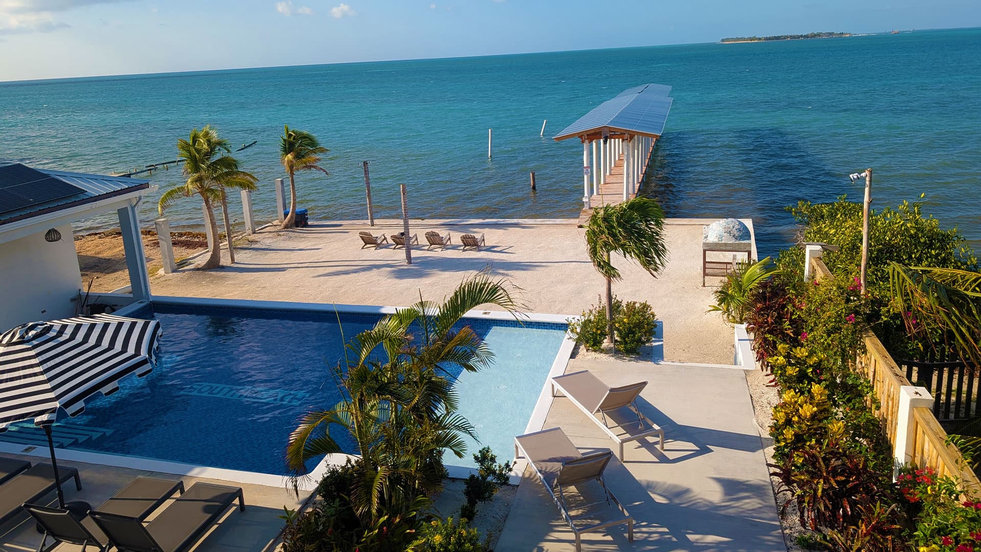 A serene beachfront view featuring a swimming pool, palm trees, and a wooden pier extending into the turquoise ocean.